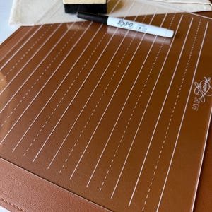 May include: A clear acrylic writing guide with vertical white dashed lines placed over a brown leather mat. The set includes a black Expo dry-erase marker, a small wooden block eraser with a black felt bottom, and a beige fabric drawstring bag.