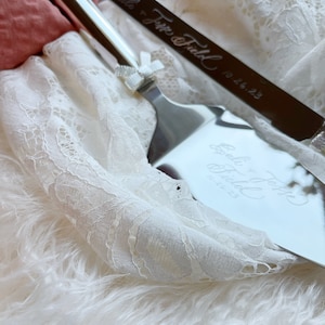 May include: A silver cake knife and server set with the inscription "So the Journey Begins" and the date "10.26.23". The set is on a white lace fabric with a white fluffy surface underneath.