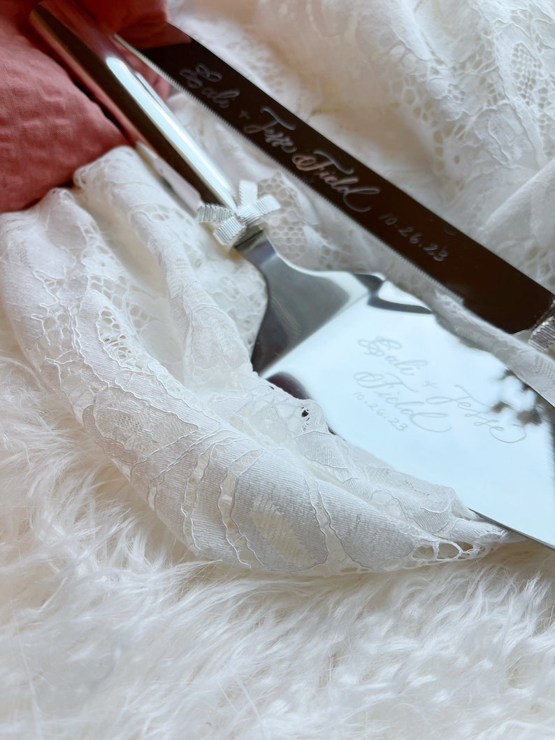 May include: A silver cake knife and server set with the inscription "So the Journey Begins" and the date "10.26.23". The set is on a white lace fabric with a white fluffy surface underneath.