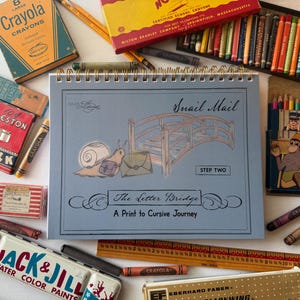 May include: A spiral-bound workbook titled The Letter Bridge: A Print to Cursive Journey, featuring a snail and bridge illustration, surrounded by vintage art supplies including Crayola crayons, Jack & Jill paints, chalk, and a wooden ruler.