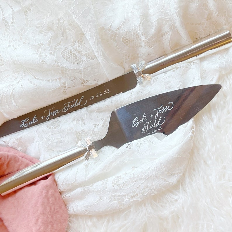 May include: A silver cake knife and server set with engraved calligraphy. The knife has the text "Gali + Jesse Field 10.26.23" and the server has the text "Gali + Jesse Field 10.26.23".