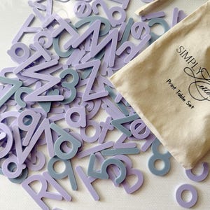 May include: A collection of light purple and muted blue plastic letters and numbers scattered alongside a beige cotton drawstring storage bag. The bag features the black printed text Simply Letters Print Table Set on its front surface.