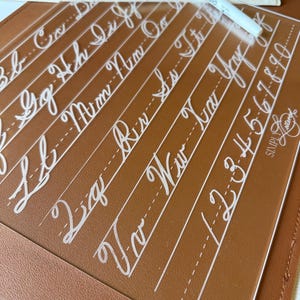 May include: A clear acrylic practice board for calligraphy featuring white printed cursive letters and numbers, accompanied by a black and white dry-erase marker and a small wooden eraser block, all resting on a brown leather-like mat.