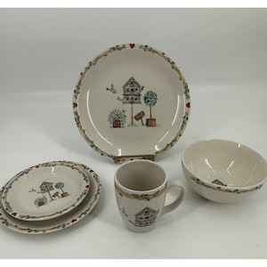 Könnte beinhalten: Ein Set cremefarbener Keramikgeschirr mit einem Vogelhaus-Design. Das Set enthält einen großen Speiseteller, einen kleineren Salatteller, eine Schüssel und eine Tasse. Das Geschirr ist mit einem grünen und braunen Blattmotiv mit roten Herzen verziert.