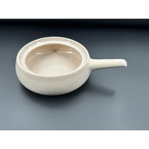 May include: A small, off-white ceramic bowl with a handle. The bowl is round with a slightly flared rim and a smooth, glossy finish. The handle is a simple, straight design. The bowl is empty and sits on a dark gray surface.