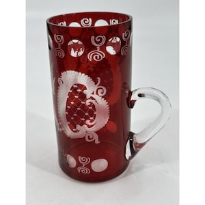 May include: A ruby red glass mug with a clear handle. The mug features white etched designs, including swirls and a central ornate pattern. The glass has a cylindrical shape and is suitable for beverages.