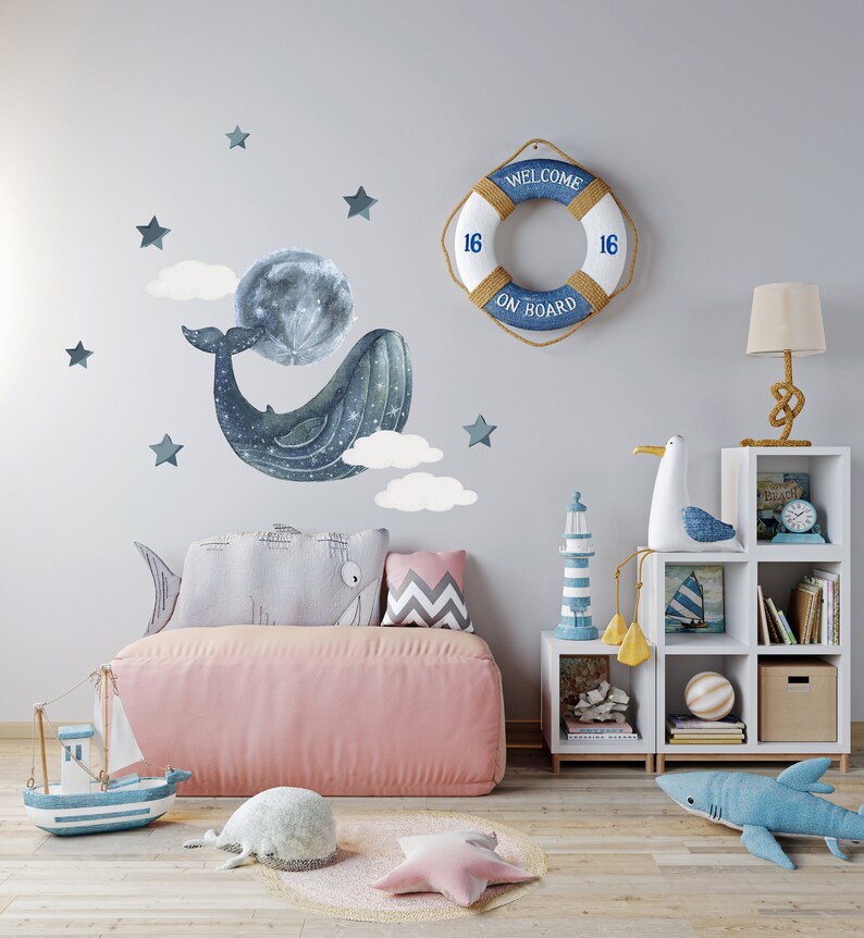 Pu&ograve; includere: Una balena blu chiaro con nuvole bianche e stelle su un muro grigio chiaro. Un salvagente con il testo "Welcome On Board" e il numero "16" su ogni lato &egrave; appeso al muro. Una libreria bianca con una statuetta di gabbiano, una statuetta di faro e libri si trova nell'angolo della stanza. Un divano rosa con un cuscino a forma di balena grigia e un cuscino grigio e bianco si trova di fronte al muro. Un tappeto bianco con un cuscino a forma di stella rosa e un cuscino a forma di balena grigia si trova sul pavimento.
