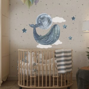 Pu&ograve; includere: Un lettino in legno con una coperta a righe bianche e grigie e una balena blu con una luna e stelle sul muro dietro.