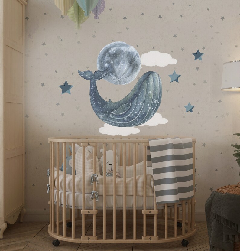 Pu&ograve; includere: Un lettino in legno con una coperta a righe bianche e grigie e una balena blu con una luna e stelle sul muro dietro.