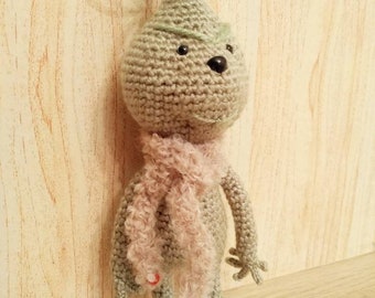 May include: A handmade crochet doll in the shape of a bulbous creature. The doll is primarily gray with a light green top and a pink scarf. It has small black eyes and a black nose. The doll is standing on a wooden surface.