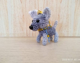 May include: A crocheted dog keychain in shades of gray and blue. The handmade dog has black eyes, a black nose, and a gold-colored collar. A blue and gold cord is attached to the collar. The dog is standing on a light brown surface.
