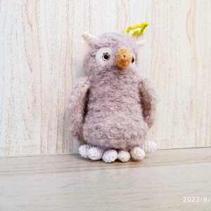 May include: A handmade, plush owl toy in shades of lavender and white. The owl has a textured, fuzzy body, white accents, and a tan beak. It has small, white feet and yellow details on its head. The toy is standing on a wooden surface.