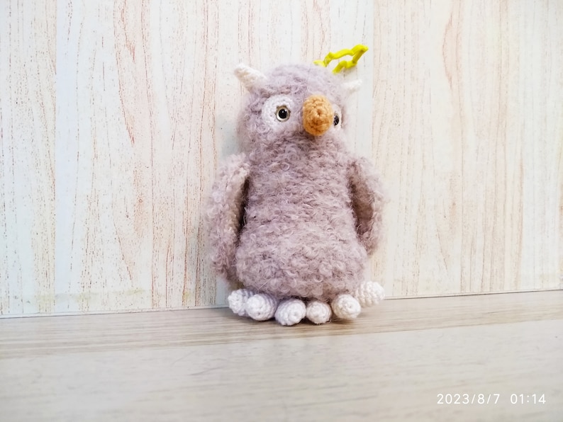May include: A handmade, plush owl toy in shades of lavender and white. The owl has a textured, fuzzy body, white accents, and a tan beak. It has small, white feet and yellow details on its head. The toy is standing on a wooden surface.