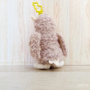 May include: A handmade, plush penguin key chain in shades of mauve and cream. The penguin has a yellow key ring and a white beak. The penguin's feet have a decorative, lacy trim. The background is a light wood.