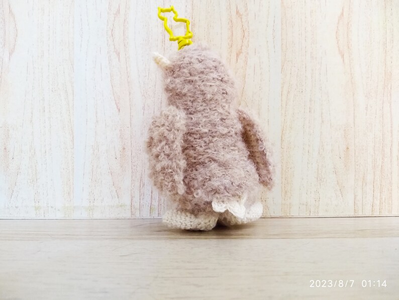 May include: A handmade, plush penguin key chain in shades of mauve and cream. The penguin has a yellow key ring and a white beak. The penguin's feet have a decorative, lacy trim. The background is a light wood.