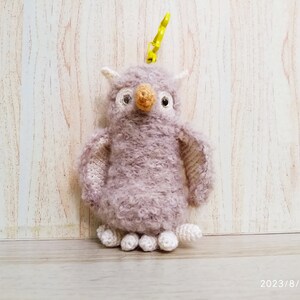 May include: A handmade, lavender-colored owl keychain with a yellow clip. The owl is crocheted with fluffy yarn, featuring white accents on the ears and feet, and a tan beak. The owl's eyes are black.