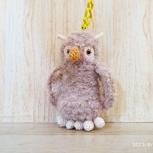 May include: A handmade, plush owl keychain in shades of lavender and white. The owl has a textured body, a tan beak, and large, round eyes. It is attached to a yellow plastic clip.