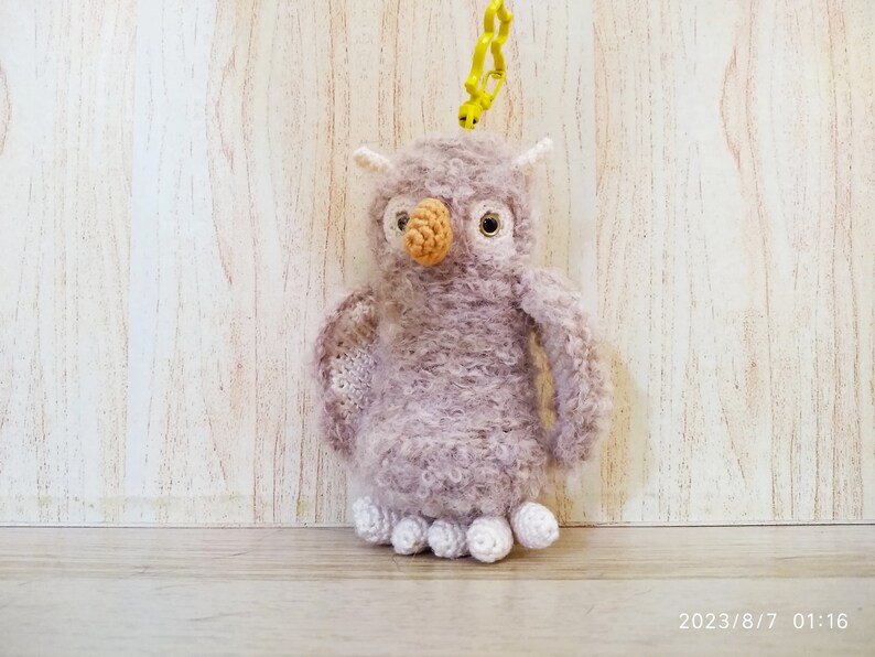 May include: A handmade, plush owl keychain in shades of lavender and white. The owl has a textured body, a tan beak, and large, round eyes. It is attached to a yellow plastic clip.