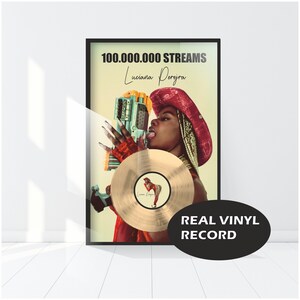 Peut inclure: Une plaque de disque d'or avec le texte "100 000 000 STREAMS" et "Luciana Perez" en texte noir. La plaque représente une femme portant un chapeau de cow-boy rouge tenant un pistolet jouet et un disque d'or. Le texte "REAL VINYL RECORD" est dans un ovale noir en bas de la plaque.