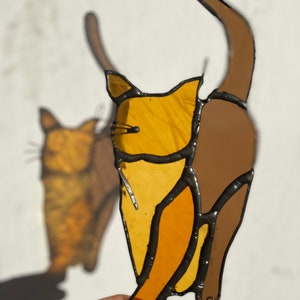 May include: Two stained glass cats, one brown and one orange, are standing side by side. The cats are outlined in black and have a simple design.