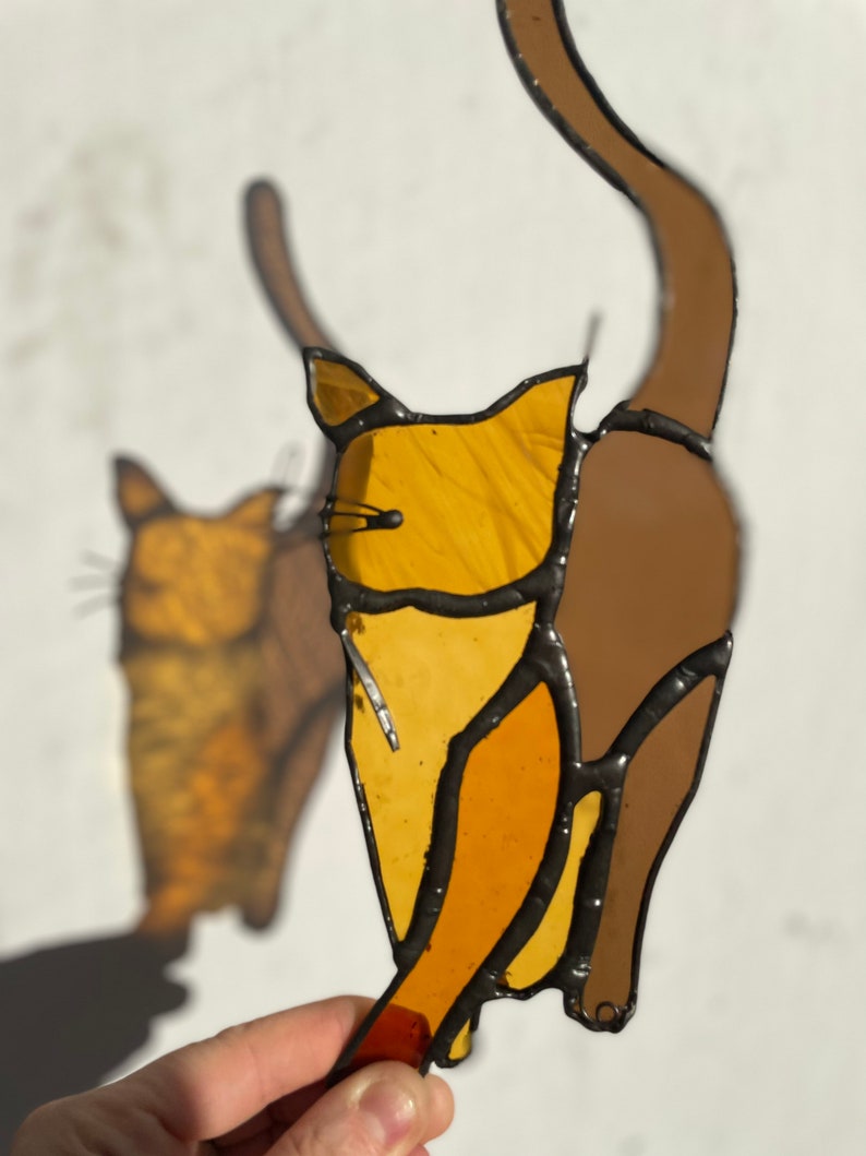 May include: Two stained glass cats, one brown and one orange, are standing side by side. The cats are outlined in black and have a simple design.