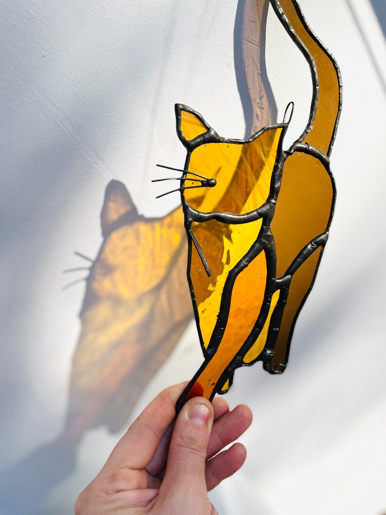 May include: A stained glass cat figurine with a yellow and orange color scheme. The cat is depicted with its tail raised in the air.