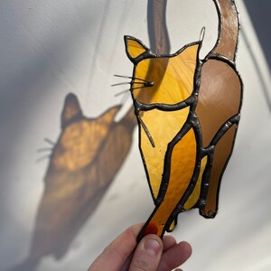 May include: A stained glass cat figurine with a brown tail and a yellow body. The cat is hanging from a string and has a shadow cast on the wall behind it.
