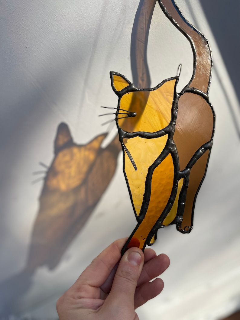 May include: A stained glass cat figurine with a brown tail and a yellow body. The cat is hanging from a string and has a shadow cast on the wall behind it.
