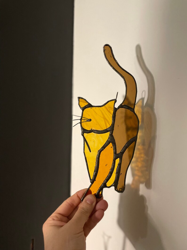 May include: A stained glass cat figurine with a yellow and brown color scheme. The cat is in a sitting position with its tail raised in the air.