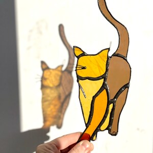 May include: A stained glass cat figurine with a brown tail and a yellow body. The cat is facing to the right and has its tail curled up behind it.