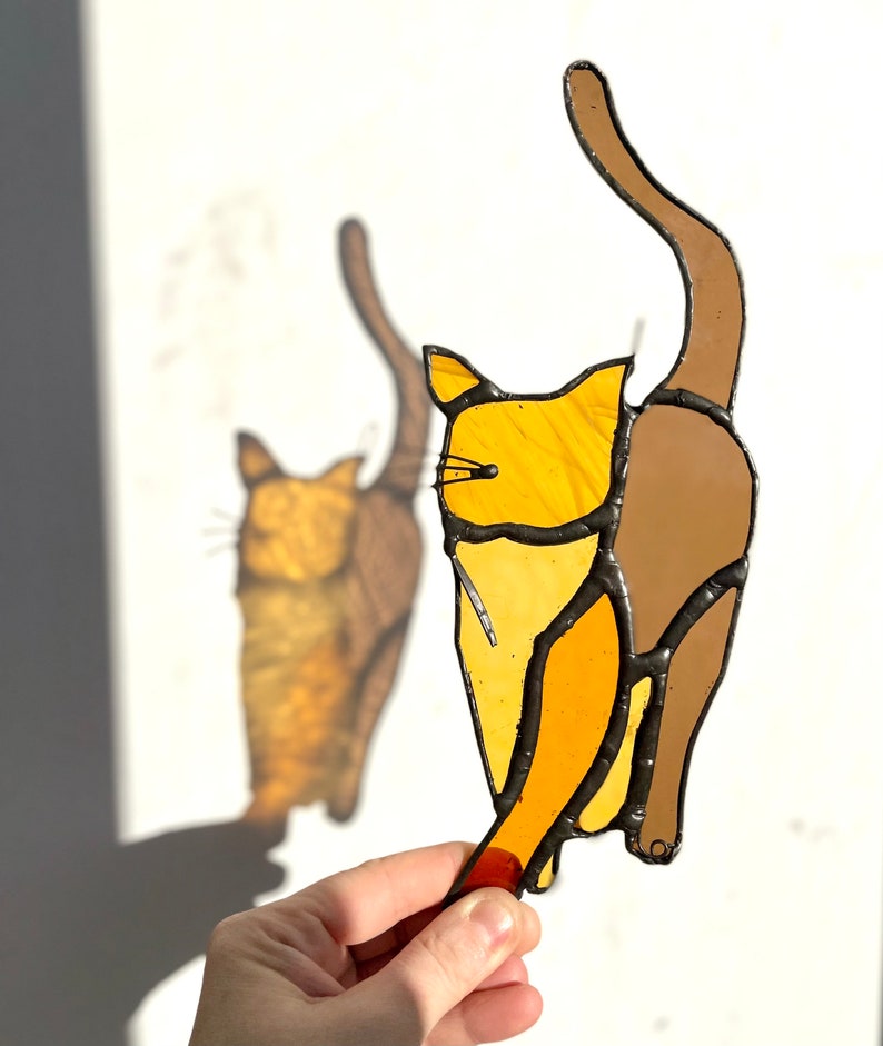 May include: A stained glass cat figurine with a brown tail and a yellow body. The cat is facing to the right and has its tail curled up behind it.