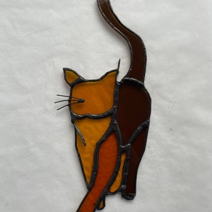 May include: A stained glass cat figurine with a brown tail and orange and yellow body.