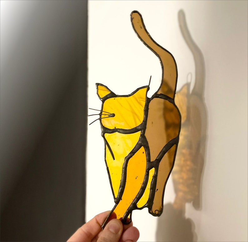 May include: A stained glass cat figurine with a yellow and brown color scheme. The cat is depicted in a playful pose, with its tail raised and its head tilted to the side.