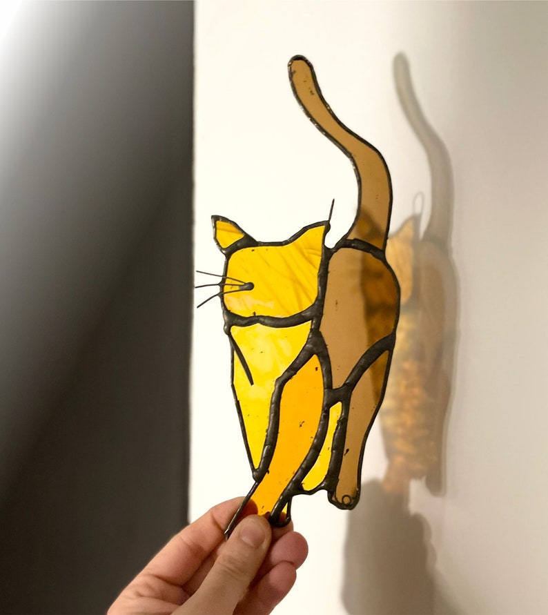 May include: A stained glass cat figurine with a yellow and brown color scheme. The cat is depicted in a playful pose with its tail raised in the air.
