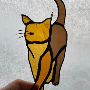 May include: A stained glass cat figurine with a brown tail and a yellow body. The cat is in a standing position with its tail curled around its body.