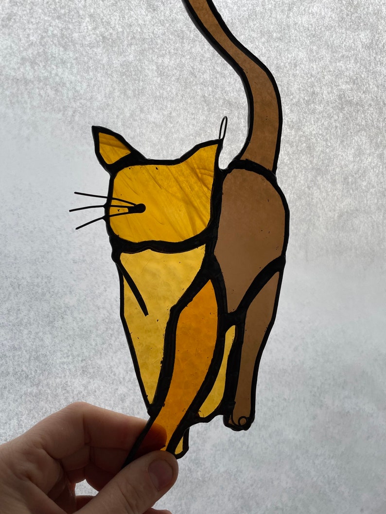 May include: A stained glass cat figurine with a brown tail and a yellow body. The cat is in a standing position with its tail curled around its body.