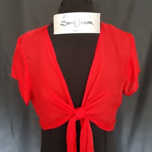 May include: A red sheer wrap top with short sleeves and a tie at the front. The wrap top is on a black mannequin.
