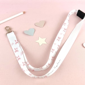 May include: A white fabric lanyard featuring a repeating pattern of small pink bows and hearts. The lanyard includes a silver-toned metal swivel clasp, a black plastic breakaway safety buckle, and the printed text "Miss Taylor" in black script.