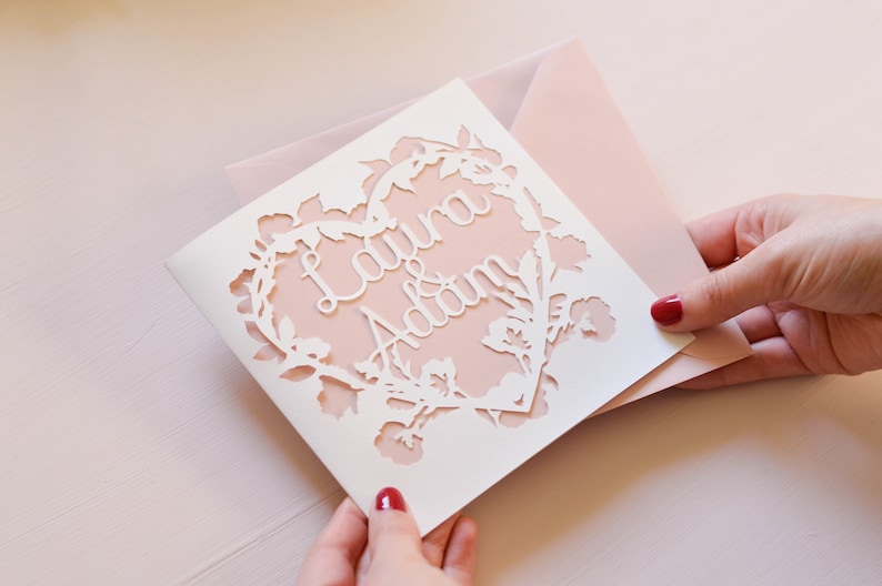 Op de afbeelding: Een witte kaart met een delicaat bloemmotief dat rond de randen is uitgesneden. Het ontwerp bevat een hartvorm met de namen "Laura & Adam" erin. De kaart is gelegd op een lichtroze kaart.