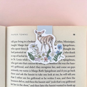 May include: A bookmark featuring a watercolour illustration of a deer standing in a field of wildflowers. The deer is brown with white spots and has large, brown eyes. The wildflowers are a variety of colours, including pink, purple, blue, and yellow. The bookmark is printed on a white background and has a blue border.