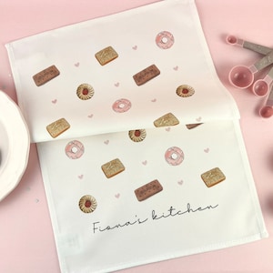 May include: White kitchen towel featuring a pattern of biscuits, doughnuts, and hearts. The towel is printed with the text "Fiana's kitchen". Measuring spoons and eggs are visible in the background.