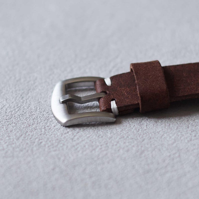 May include: Close-up of a brown leather watch strap with a silver-colored buckle. The strap is made of a textured, suede-like material. White stitching is visible near the buckle. The buckle is rectangular with rounded edges.