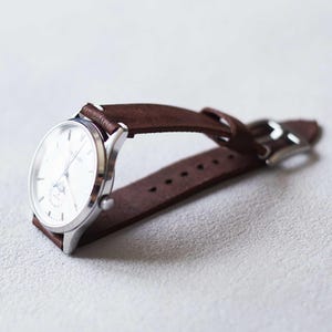 May include: A silver wristwatch with a white face and brown leather band. The watch has a classic design with a small subdial and a buckle closure. The band is made of a soft, suede-like material.