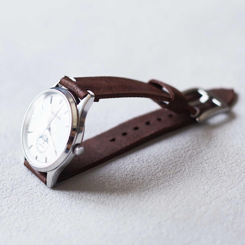 May include: A silver wristwatch with a white face and brown leather band. The watch has a classic design with a small subdial and a buckle closure. The band is made of a soft, suede-like material.