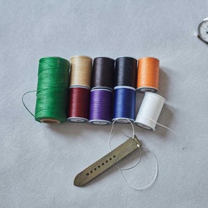 May include: A collection of colorful spools of thread in green, tan, brown, black, orange, white, red, purple, and blue. A leather watch strap is positioned below the spools, with a needle and thread attached, suggesting a craft or repair project.