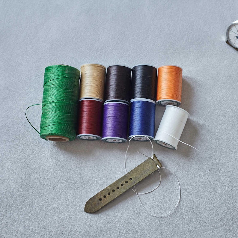 May include: A collection of colorful spools of thread in green, tan, brown, black, orange, white, red, purple, and blue. A leather watch strap is positioned below the spools, with a needle and thread attached, suggesting a craft or repair project.