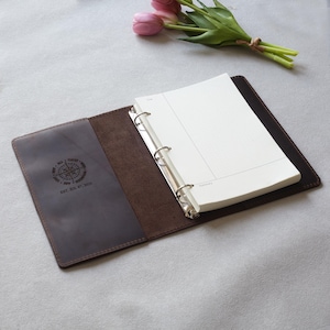 Puede incluir: Una carpeta de cuero marrón oscuro con un mecanismo de tres anillas de metal y papel cuadriculado blanco. El panel interior izquierdo tiene grabado un diseño de brújula circular y el texto EST. JUL 07, 2026. Un pequeño ramo de tulipanes rosas.