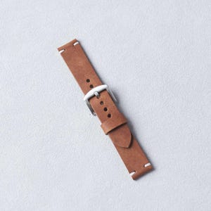 May include: A brown leather watch strap with a silver buckle. The strap has white stitching at the ends and multiple holes for adjustment. The leather has a textured appearance.