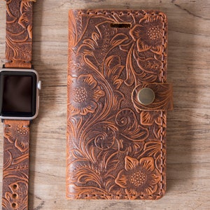 May include: A brown leather Apple Watch band and phone case with a floral embossed design. The watch band is connected to a silver Apple Watch. The phone case has a snap closure.