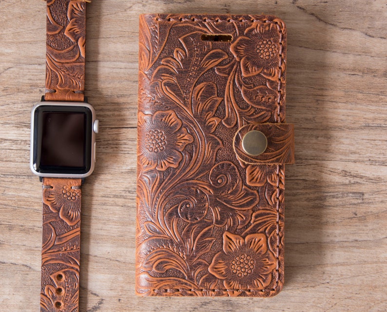 May include: A brown leather Apple Watch band and phone case with a floral embossed design. The watch band is connected to a silver Apple Watch. The phone case has a snap closure.